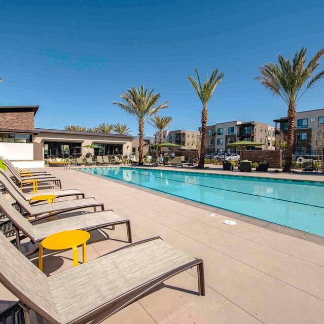 Redlands, CA Apartments - The Summit - Sparkling Swimming Pool Surrounded by Lounge Seating, Side Tables, and Palm Trees. Views of the Clubhouse, and Apartments in Background
