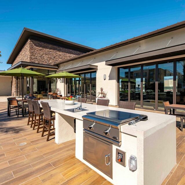 Apartments Redlands, CA - The Summit - Outdoor BBQ and Picnic Area with Chairs, Tables, Umbrellas, Kitchen Area, and Clubhouse Lounge in Background